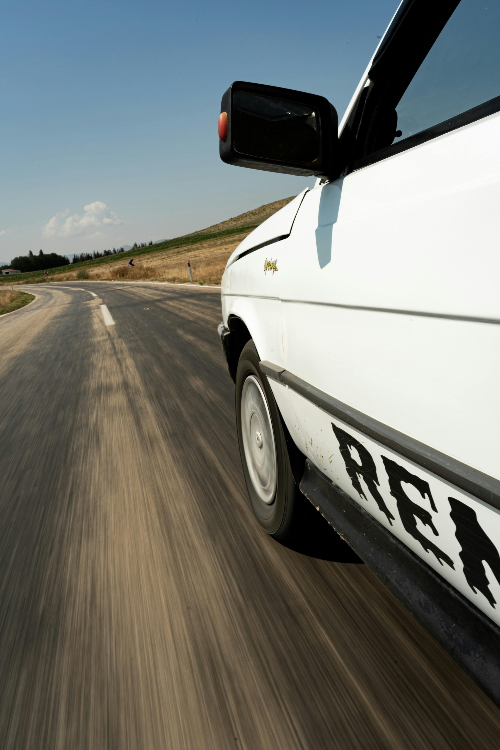 View of a vintage white car speeding down a winding rural road under a clear sky.