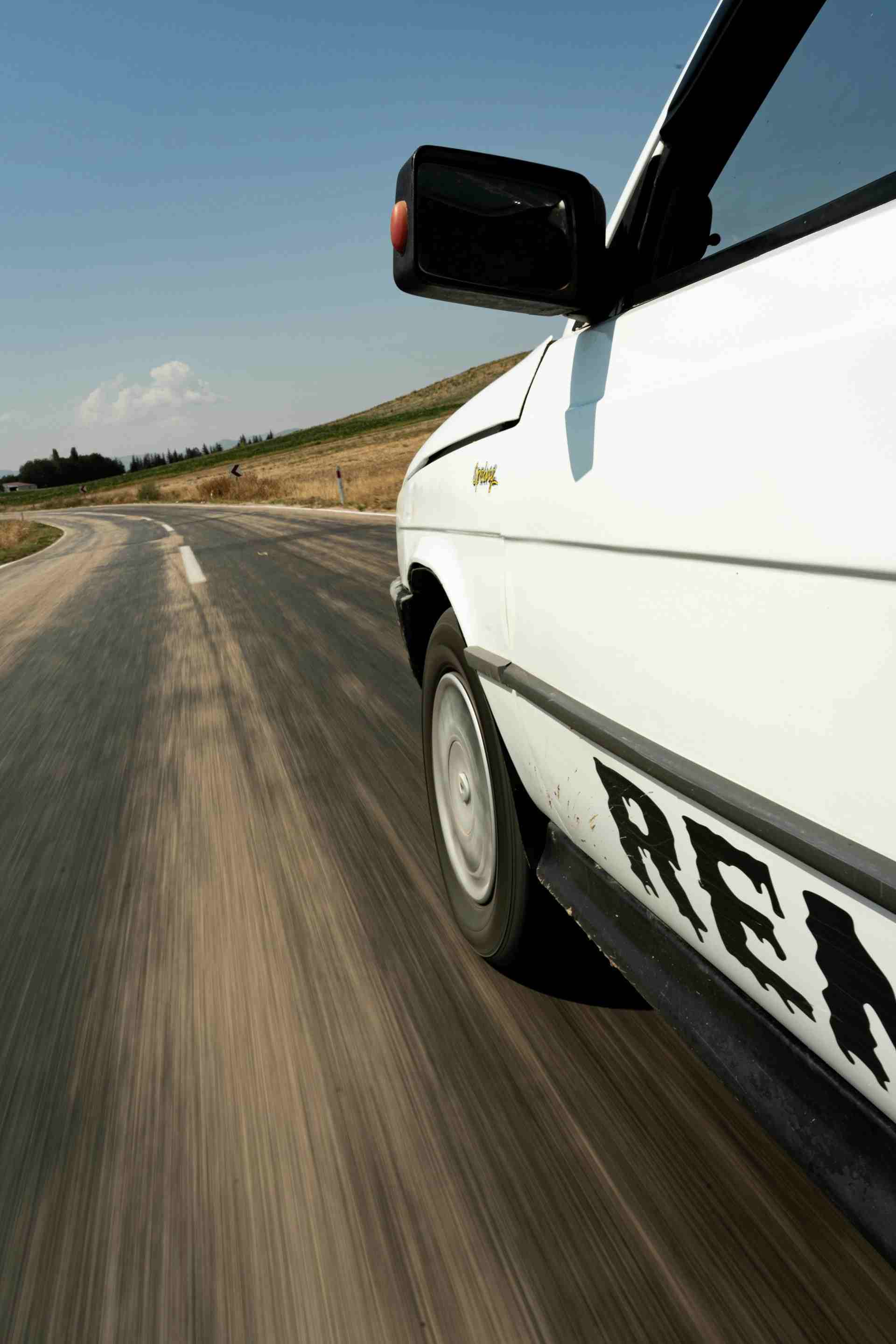 View of a vintage white car speeding down a winding rural road under a clear sky.